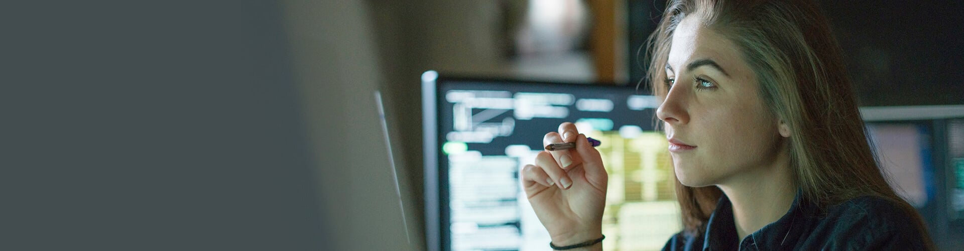 The woman is holding a pencil in her hand and is analyzing something on the monitor