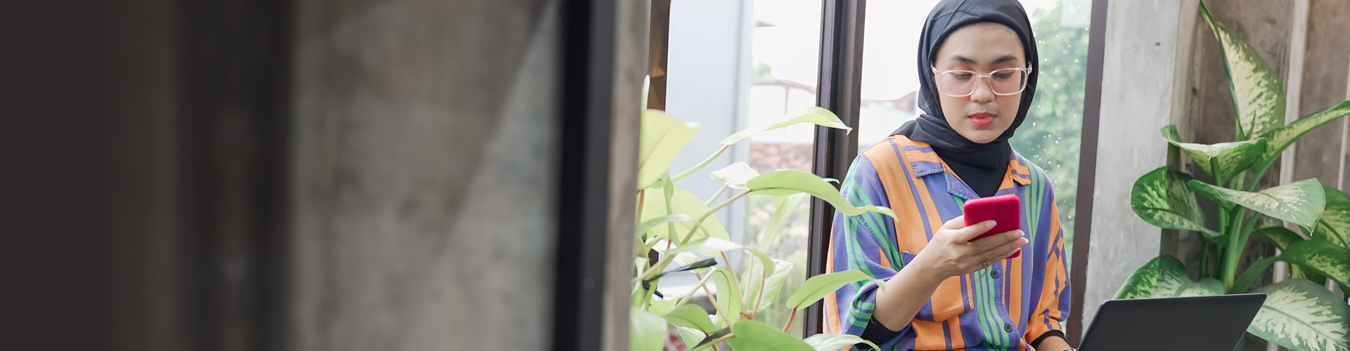 Muslim young lady leaning against a windowns in between two plants looking at a mobile phone screen