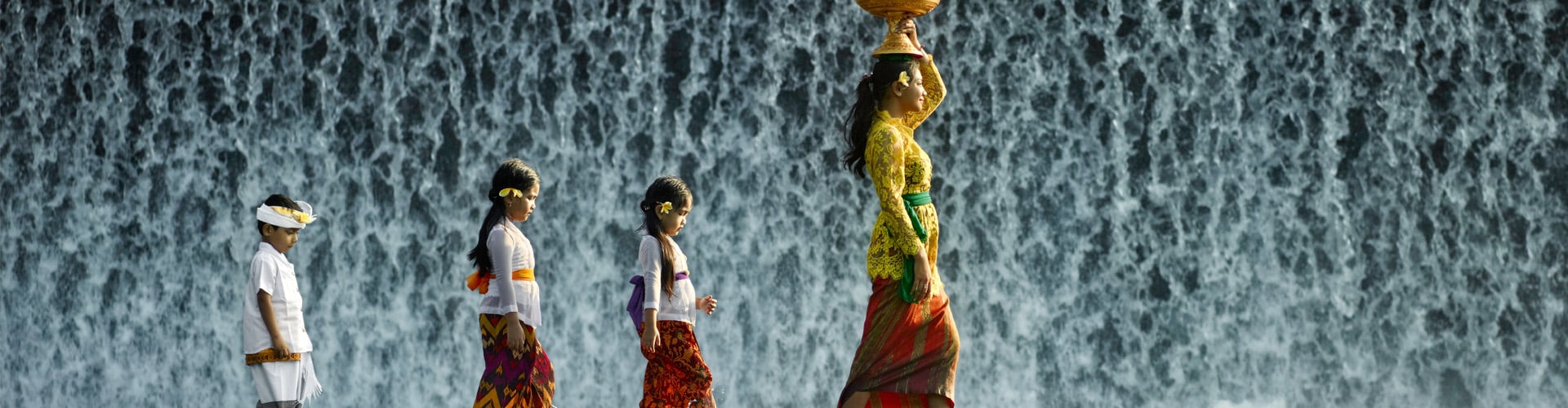 Young Balinese woman and children wearing traditional clothing, walking across waterfall
