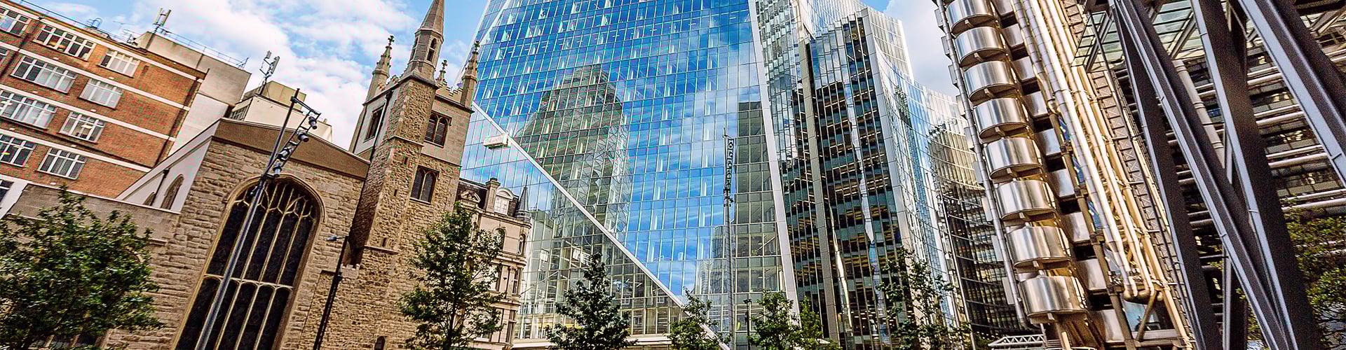 Cityscape showing modern glass skyscrapers reflecting the sky, alongside an older stone church and contemporary steel structures in an urban setting.