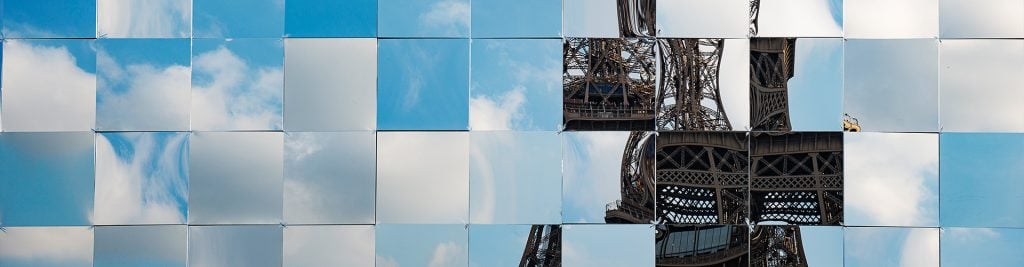 A collage of square mirrors reflecting the sky with clouds and parts of the Eiffel Tower. The mirrors are arranged in a grid pattern, creating a fragmented and artistic view of the iconic structure against a blue sky backdrop.