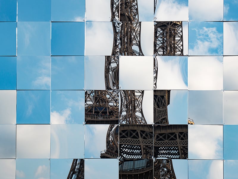 A collage of square mirrors reflecting the sky with clouds and parts of the Eiffel Tower. The mirrors are arranged in a grid pattern, creating a fragmented and artistic view of the iconic structure against a blue sky backdrop.