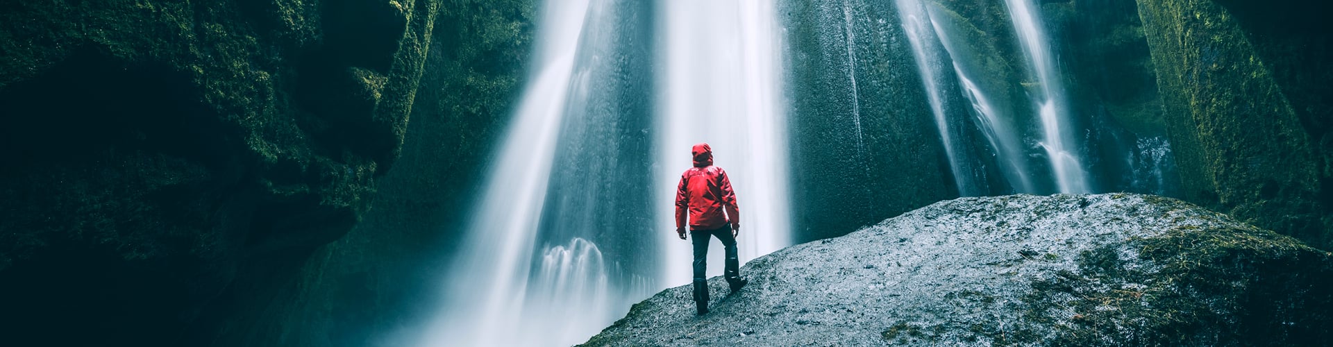 Person wearing a red jacket standing on a moss-covered rock inside a narrow canyon, looking at tall, cascading waterfalls surrounded by dark green cliffs