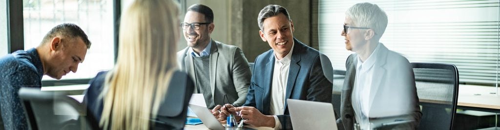 Group of professionals seated around a conference table in a modern office setting, engaged in discussion. Laptops, documents, and pens are visible on the table, with large windows and blinds in the background.