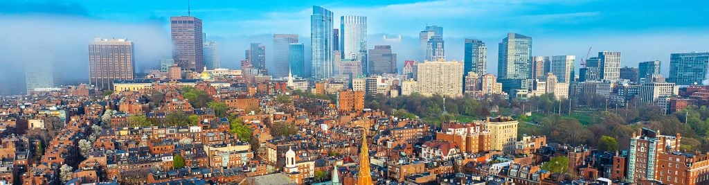 Panoramic view of a city skyline with modern glass skyscrapers in the background and historic brick buildings in the foreground, under a clear blue sky with light fog partially covering some towers.