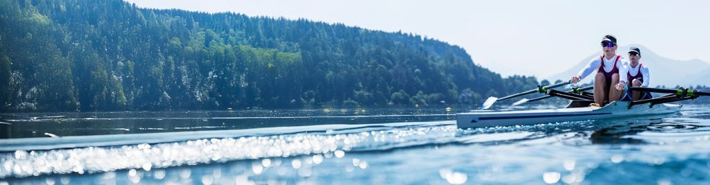 Two rowers in a racing shell glide across a calm lake, creating ripples on the water, with a forested shoreline and distant mountains in the background.