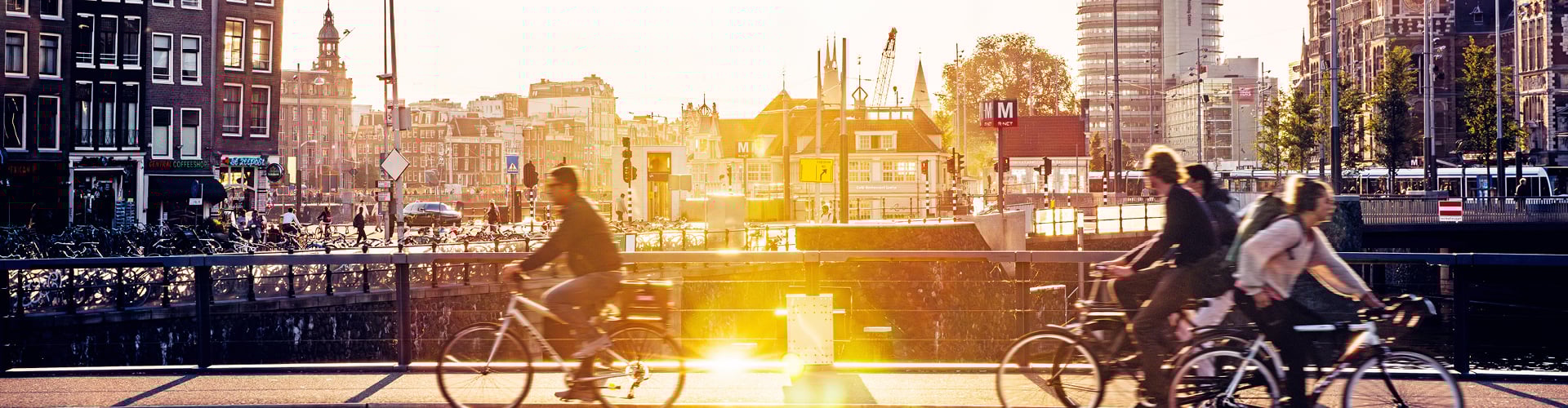 Cyclists riding along a canal-side street in Amsterdam at sunset, with historic buildings, bridges, and city infrastructure in the background.