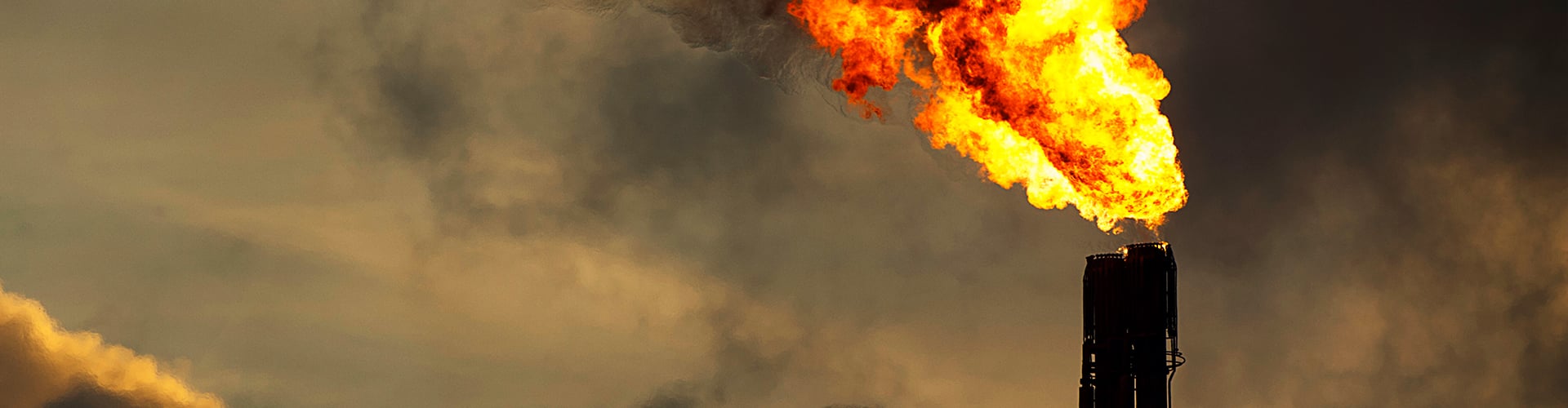 A tall industrial flare stack burning a large, bright orange flame against a cloudy sky, with dark smoke partially visible in the background.