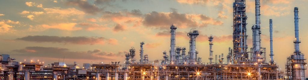 Large industrial oil and gas refinery with multiple tall distillation towers, metal piping, and illuminated structures at sunset, with a sky of warm clouds in the background.