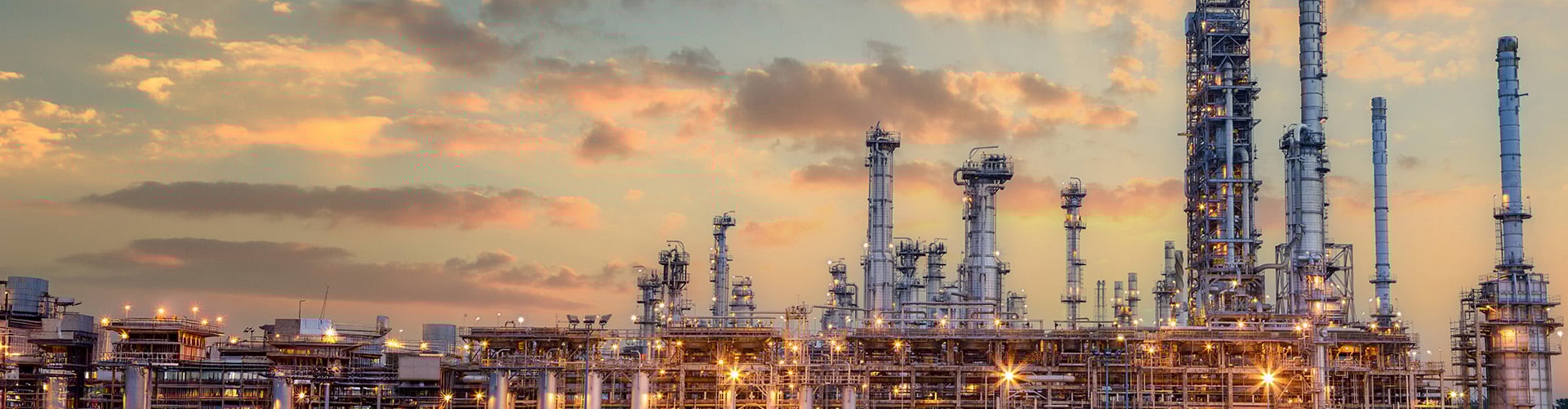 Large industrial oil and gas refinery with multiple tall distillation towers, metal piping, and illuminated structures at sunset, with a sky of warm clouds in the background.