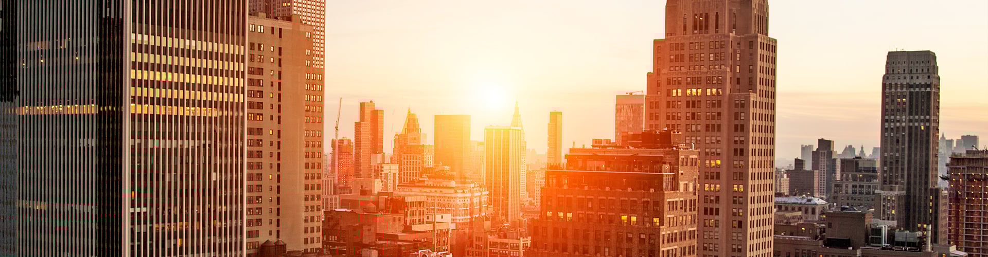 Sunset over a city skyline with tall buildings and warm orange light illuminating the scene.