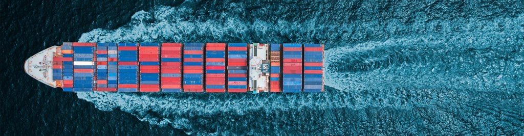Aerial, top-down view of a large container ship carrying red and blue shipping containers, moving through dark blue ocean water and leaving a white wake behind.