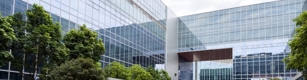 A modern glass office building with large reflective windows, surrounded by tall green trees in the foreground.