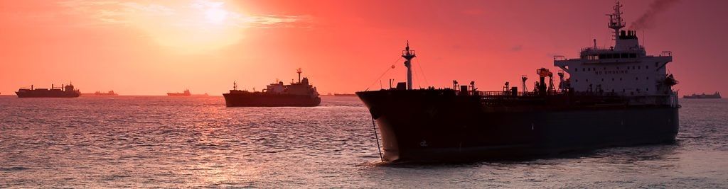 Large cargo ships sailing across calm water at sunset, with an orange‑red sky and the sun low on the horizon.