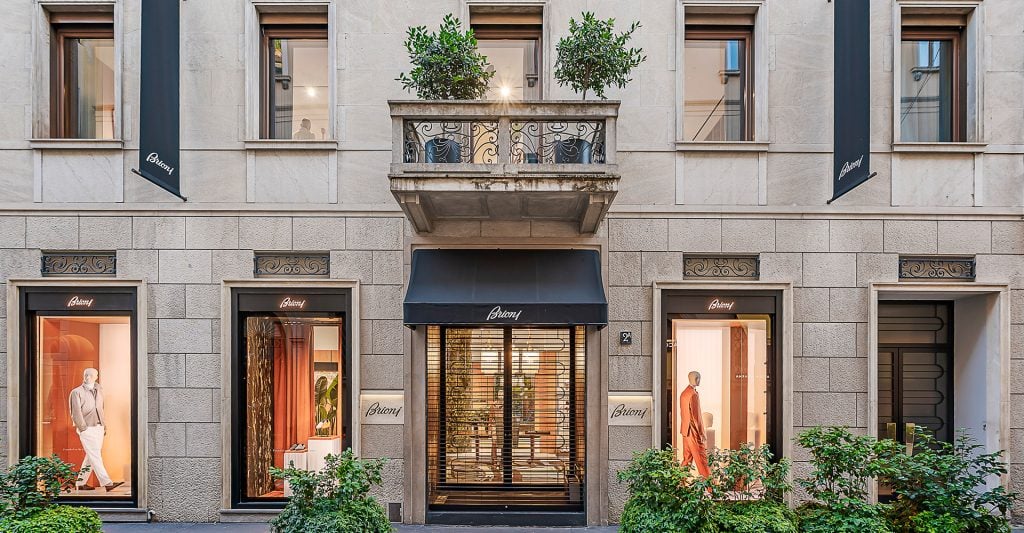 Exterior view of a luxury boutique storefront with large display windows showing mannequins dressed in elegant clothing. The building features light stone walls, decorative iron balcony with potted plants, and a central entrance with a black awning. Green shrubs line the front of the store.