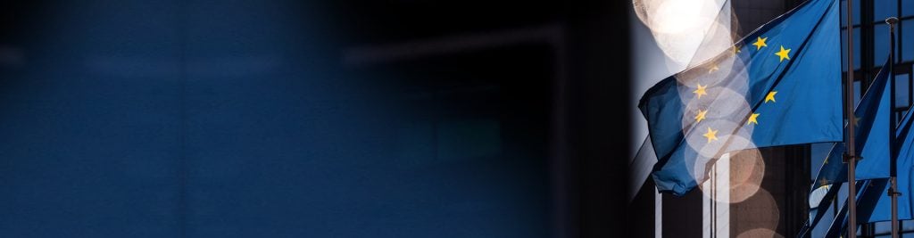 European Union flag waving outside a modern building, with light reflections and a dark blue background providing space for text.