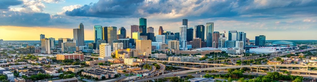 Panoramic view of a modern city skyline with high‑rise office buildings and residential towers, seen above highways and neighbourhoods in the foreground, under a partly cloudy sky at sunset.