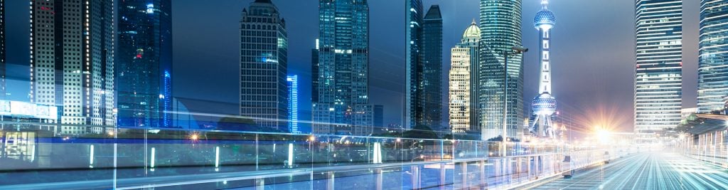 Nighttime city skyline with illuminated skyscrapers and light trails along a modern pedestrian walkway.