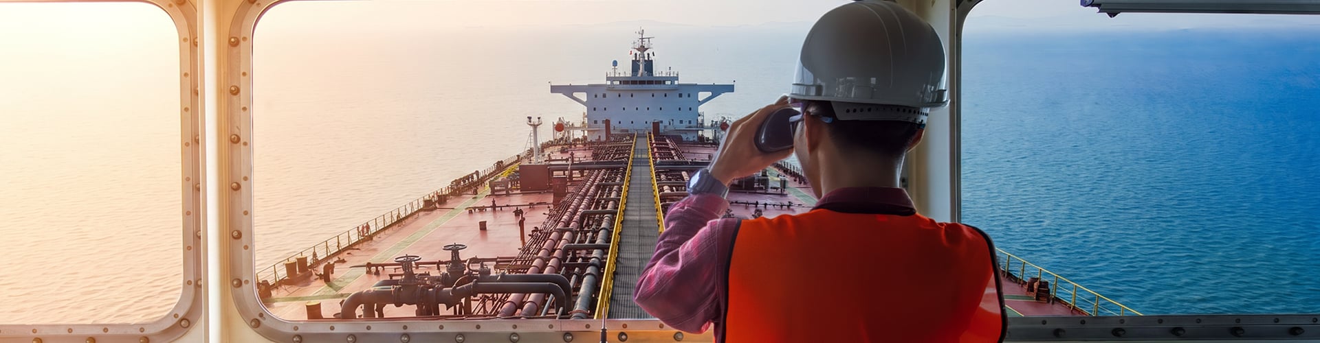 View from inside a ship’s bridge looking out over the deck and open sea, with a worker in a high-visibility orange vest and white hard hat standing in the foreground, holding binoculars. The ship’s deck is lined with pipes leading toward a large vessel structure at the front, while control panels, communication equipment, and windows frame the interior. The ocean stretches to both sides under soft, warm light.