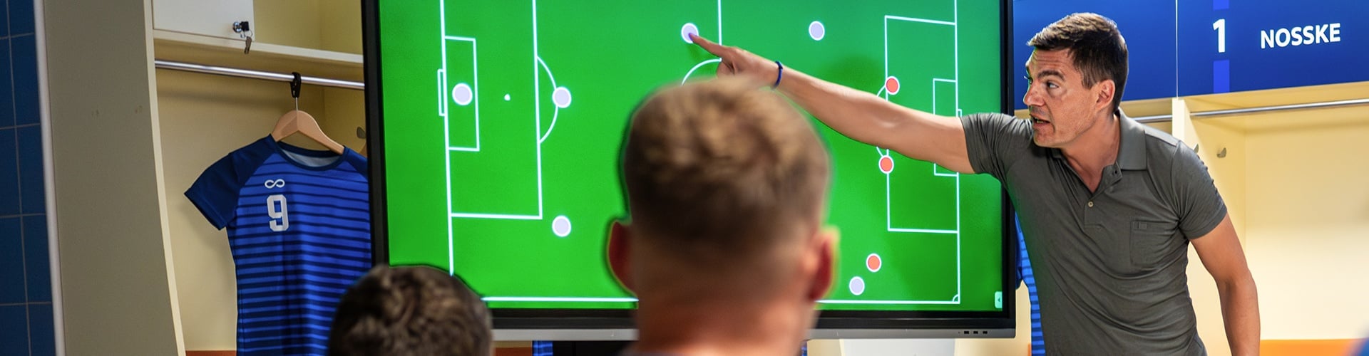 Person pointing at a football tactics screen in a changing-room setting, with young players watching from the foreground.