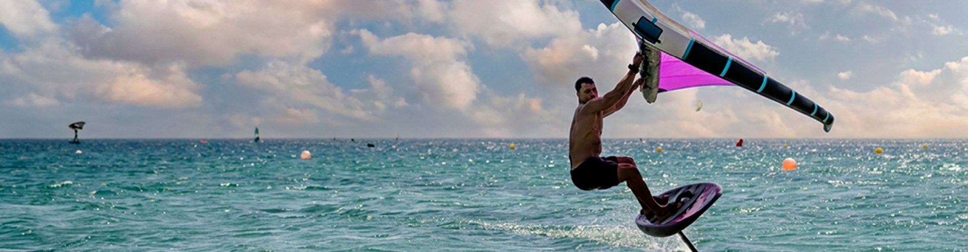 Person riding a hydrofoil board across the ocean while holding a colorful wing sail, with calm turquoise water and a cloudy sky in the background.