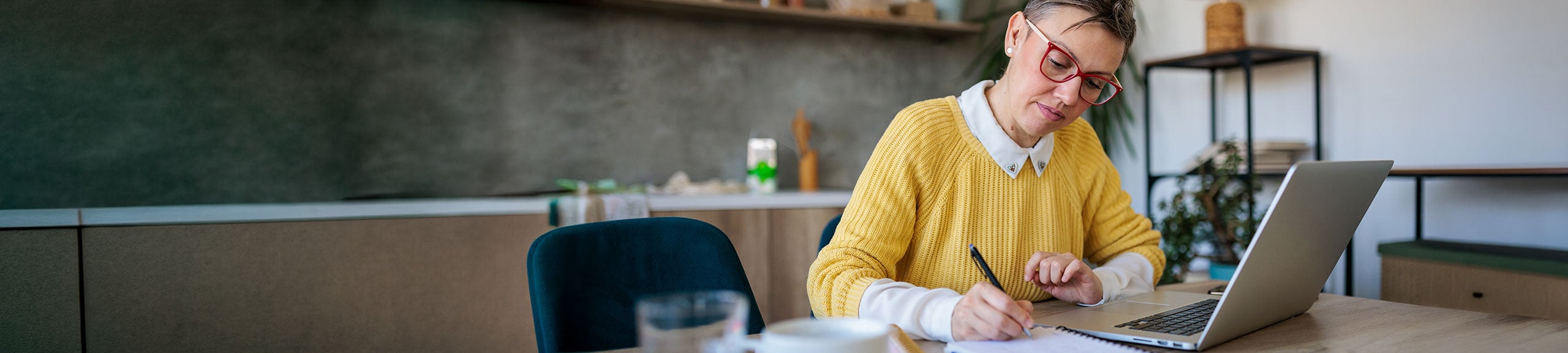 Woman wearing yellow jumper sitting in kitchen working with laptop and writing notes