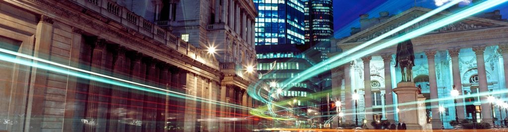 Historic and modern city buildings at night with colorful traffic light trails and illuminated streets.