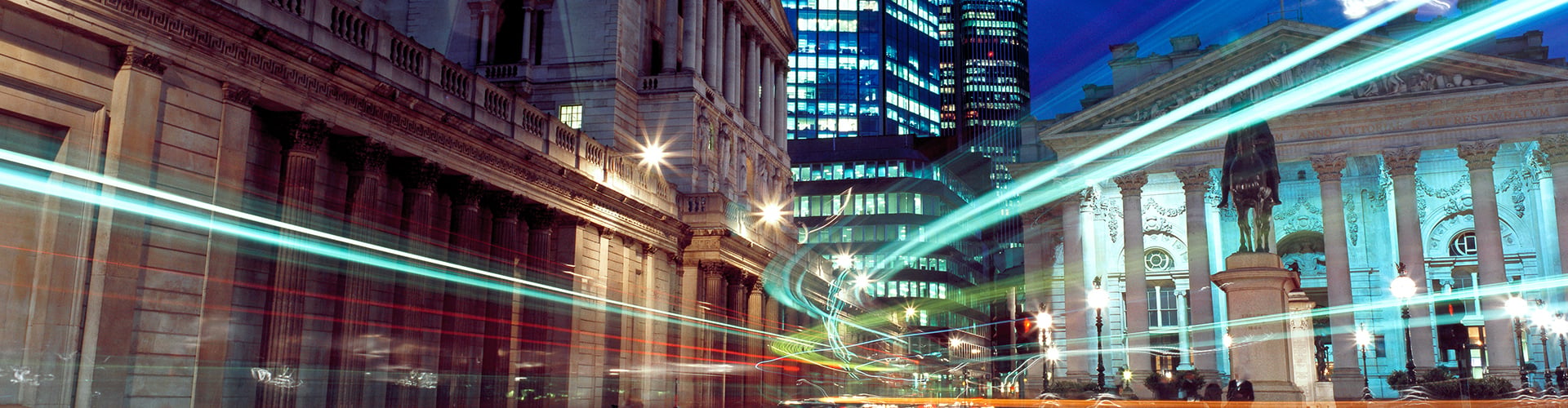 Historic and modern city buildings at night with colorful traffic light trails and illuminated streets.