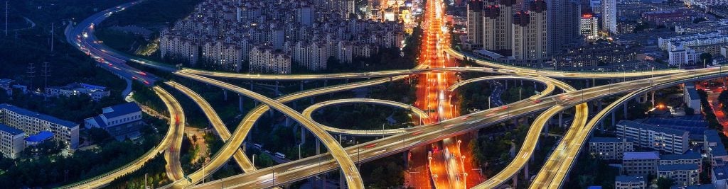 Aerial view of a brightly lit urban highway interchange at night, with multiple elevated roads weaving through a cityscape and vehicle light trails illuminating the scene.