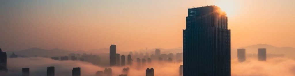 Sunrise over a modern city skyline, with a tall skyscraper illuminated by the morning sun rising above a layer of low clouds and mist.