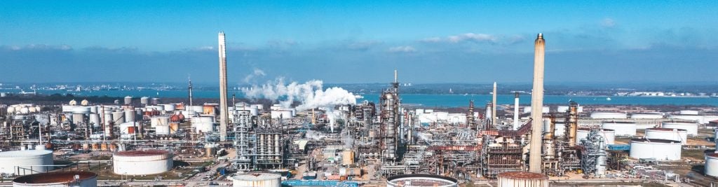 Panoramic view of a large industrial refinery with storage tanks, processing equipment, tall chimneys, and steam emissions, set against a blue sky and waterfront.