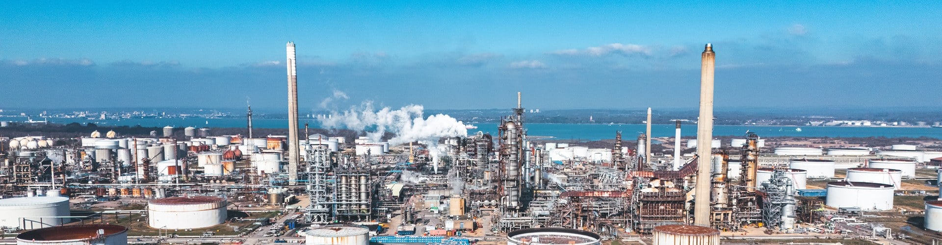 Panoramic view of a large industrial refinery with storage tanks, processing equipment, tall chimneys, and steam emissions, set against a blue sky and waterfront.