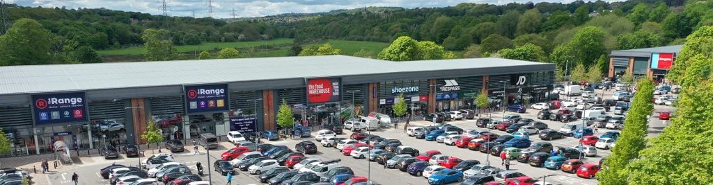 Retail park with multiple stores and a busy car park, surrounded by green woodland.