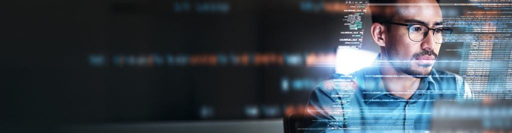 Man wearing glasses looking at laptop with financial figures on screen in the background